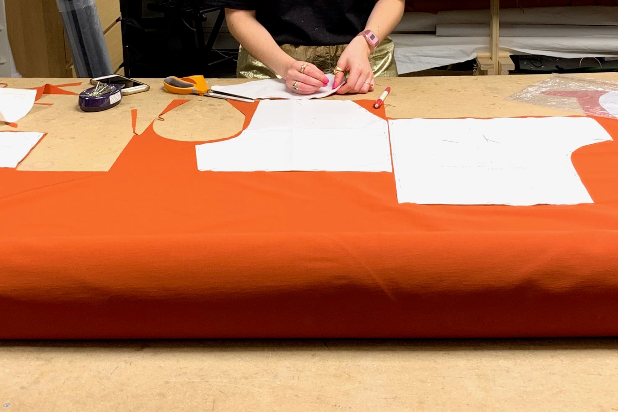 Une personne avec des ongles rose et des bijoux dorés positionnant des patrons au-dessus d'un coupon de tissu orange sur le plan de travail d'un atelier de confection